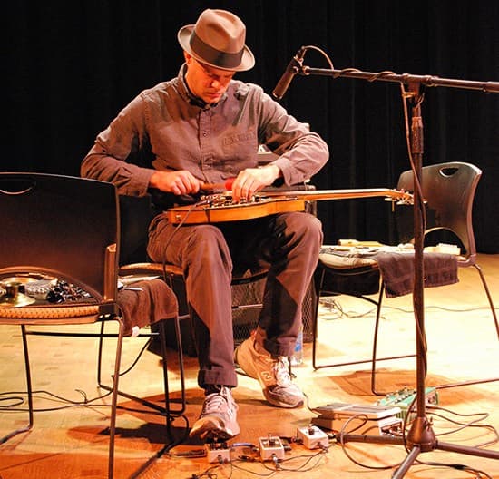 A person wearing a hat is playing a string instrument while seated on a stage, surrounded by equipment and a microphone.