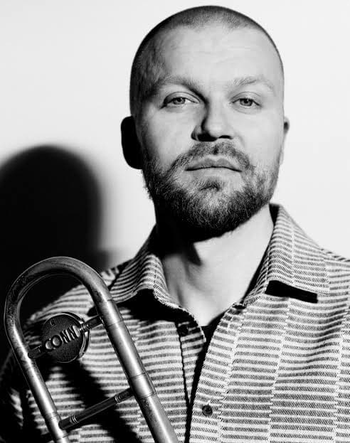 A man with a beard holds a trombone, wearing a patterned shirt, in a black and white setting.