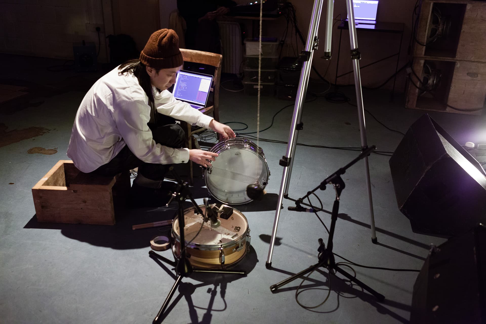 A person in a beanie adjusts a drum near audio equipment and a laptop in a studio setting.