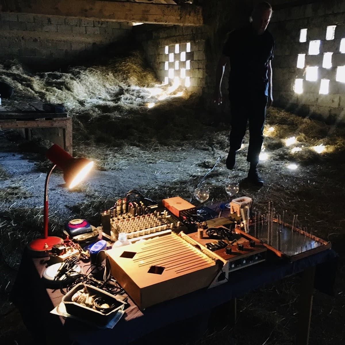 A dimly lit room with hay on the floor and a table covered with electronic music equipment, illuminated by a small lamp.