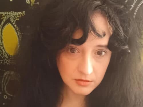 A person with long, dark, curly hair is looking directly at the camera against a patterned background.