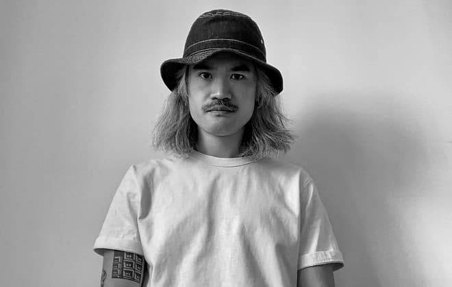 A person with long hair and a mustache wears a bucket hat and a plain t-shirt, standing against a plain background.
