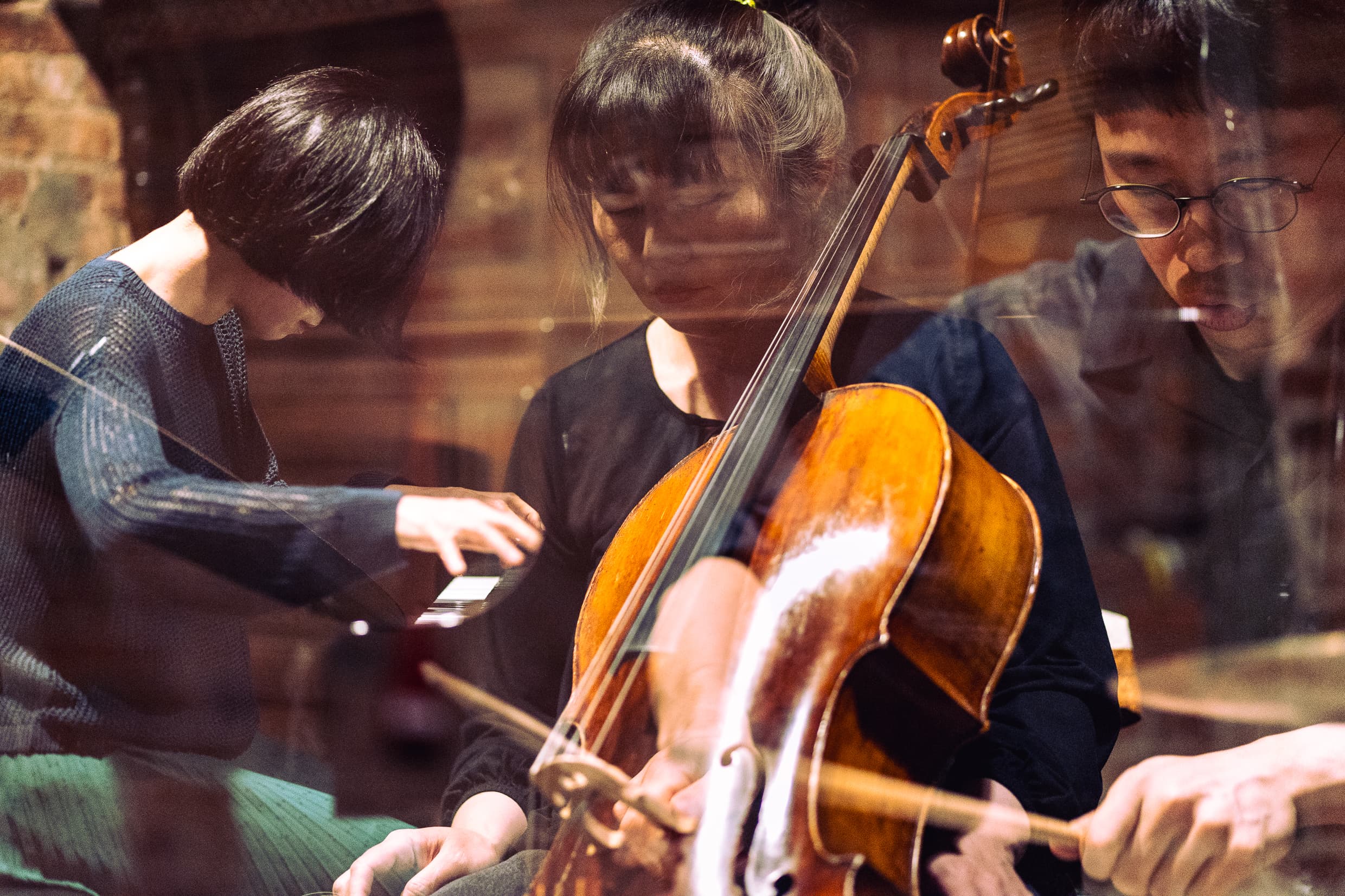 Three musicians are playing instruments, with one on a piano, another on a cello, and the third drums, all in a reflective view.
