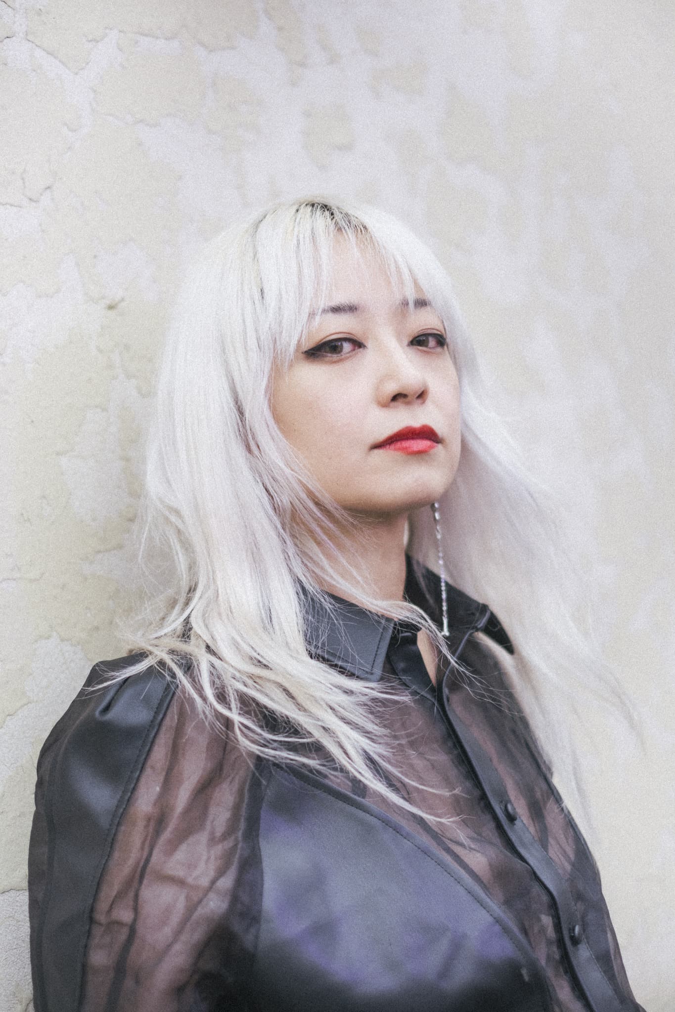A person with long white hair and red lipstick is wearing a sheer black shirt, standing against a textured wall.
